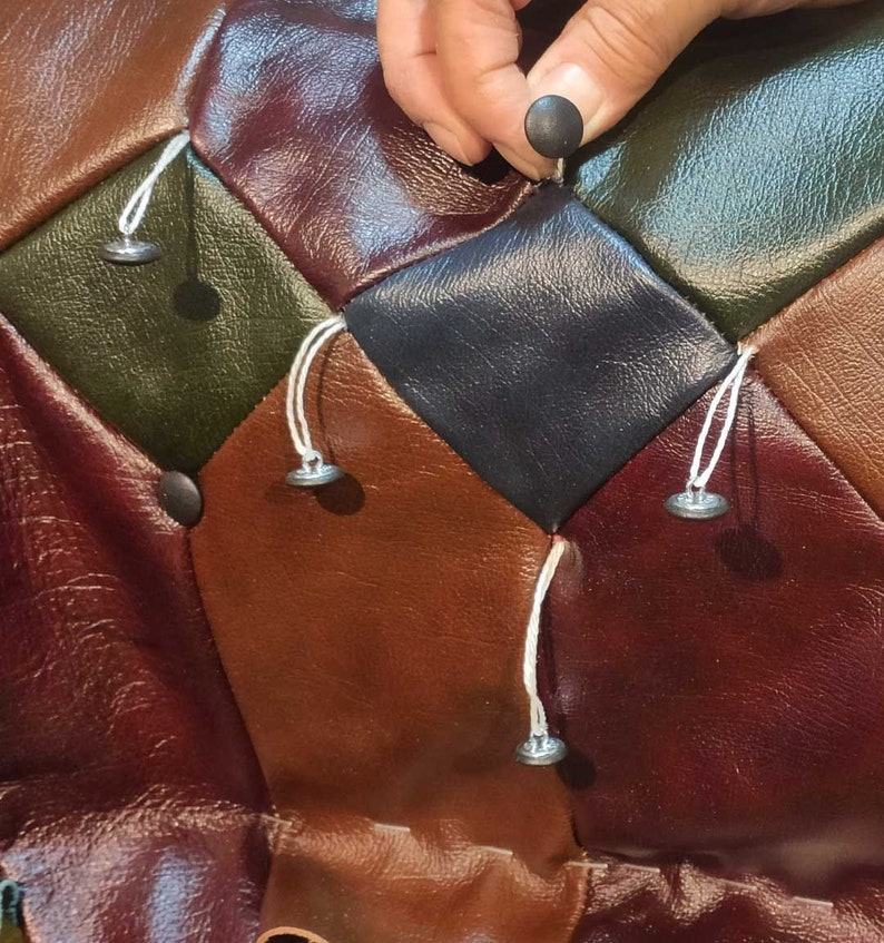 May include: Close-up of a multi-colored, diamond-patterned leather surface. The colors include shades of brown, green, and black. A hand is shown placing a black button. Silver buttons are attached with white string.