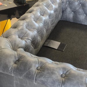 May include: A gray, tufted leather sofa with a classic Chesterfield design. The sofa features button detailing and a distressed finish, showcasing a vintage aesthetic. The image also shows a cordless drill and other tools.