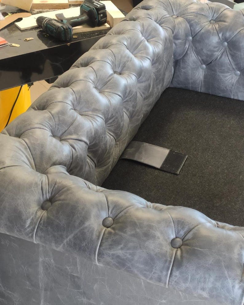 May include: A gray, tufted leather sofa with a classic Chesterfield design. The sofa features button detailing and a distressed finish, showcasing a vintage aesthetic. The image also shows a cordless drill and other tools.