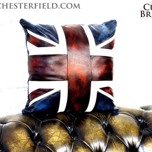 May include: A leather cushion featuring a Union Jack design. The cushion is red, white and blue and has a distressed look. The cushion is resting on a brown leather sofa.