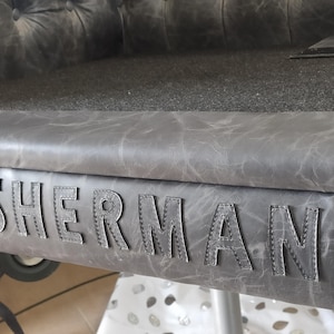 May include: Close-up of a gray leather bench with the name "SHERMAN" stitched across the front. The bench has a tufted back and a textured surface. The color is a muted gray.