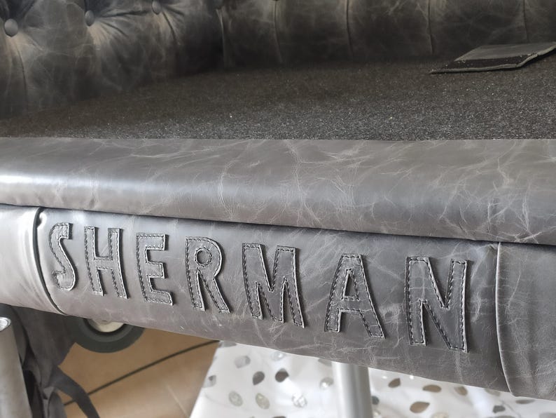 May include: Close-up of a gray leather bench with the name "SHERMAN" stitched across the front. The bench has a tufted back and a textured surface. The color is a muted gray.