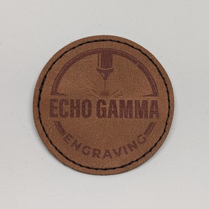 Könnte beinhalten: Ein braunes Leder-Patch mit dem Text "ECHO GAMMA ENGRAVING" in mahagonifarbener Schrift.