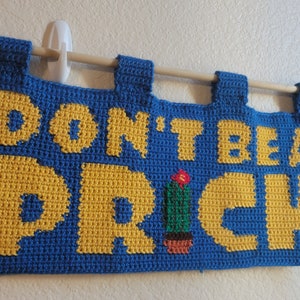 Può includere: Un arazzo da parete in crochet blu e giallo con il testo "Don't be a prick" e un piccolo cactus verde con un fiore rosso.
