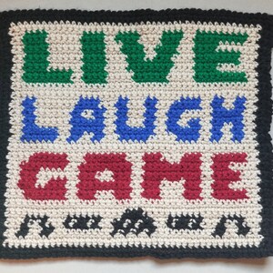 May include: Crocheted wall hanging with the words "LIVE LAUGH GAME" in green, blue, and red yarn. The background is white with a black border.
