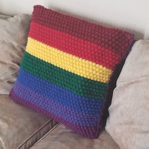 May include: A rainbow-colored crocheted pillow with a textured surface. The pillow is made of multiple stripes of maroon, red, yellow, green, blue, and purple yarn.