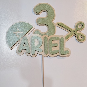 Op de afbeelding: Groene en witte taarttopper met het nummer 3, de naam "Ariel" en een schaar.