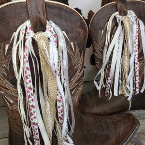 May include: A pair of dark brown leather cowboy boots featuring intricate gold stitching on the shafts and square toes. Each boot is adorned with a decorative cluster of hanging ribbons in white, tan, and a floral pattern tied to the pull tabs.