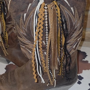 May include: A brown leather cowboy boot featuring white stitched wing patterns on the shaft and a decorative tassel attachment made of various ribbons, including gold, black, and white rickrack, braided cords, and studded leather strips.