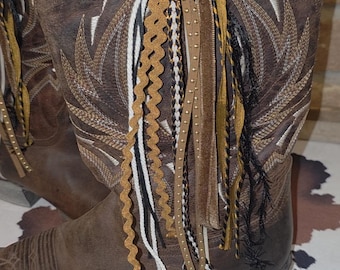 May include: A brown leather cowboy boot featuring white stitched wing patterns on the shaft and a decorative tassel attachment made of various ribbons, including gold, black, and white rickrack, braided cords, and studded leather strips.