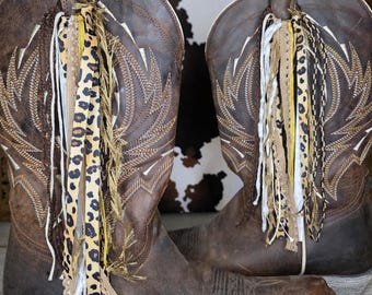 May include: A pair of brown leather cowboy boots featuring intricate white stitching on the shaft and a square toe. Each boot is adorned with a decorative tassel attachment made of various ribbons, including leopard print, frayed fabric, and gold trim.