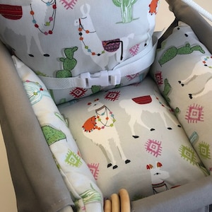 May include: A gray and white patterned high chair cushion with a white safety strap and buckle. The cushion features a llama and cactus design.