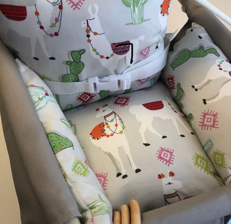 May include: A gray and white patterned high chair cushion with a white safety strap and buckle. The cushion features a llama and cactus design.