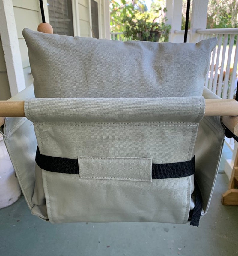 May include: A light gray canvas swing seat with a black strap and a matching light gray pillow. The swing seat is hanging from a wooden frame.