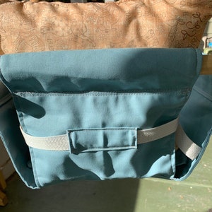 May include: A teal blue fabric swing seat with a white strap and a pocket. The seat is attached to a wooden frame.