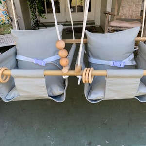 May include: Two gray canvas swing seats with wooden bars and rope hanging straps. Each seat has a gray cushion and a purple buckle strap. There are wooden beads on the bar.