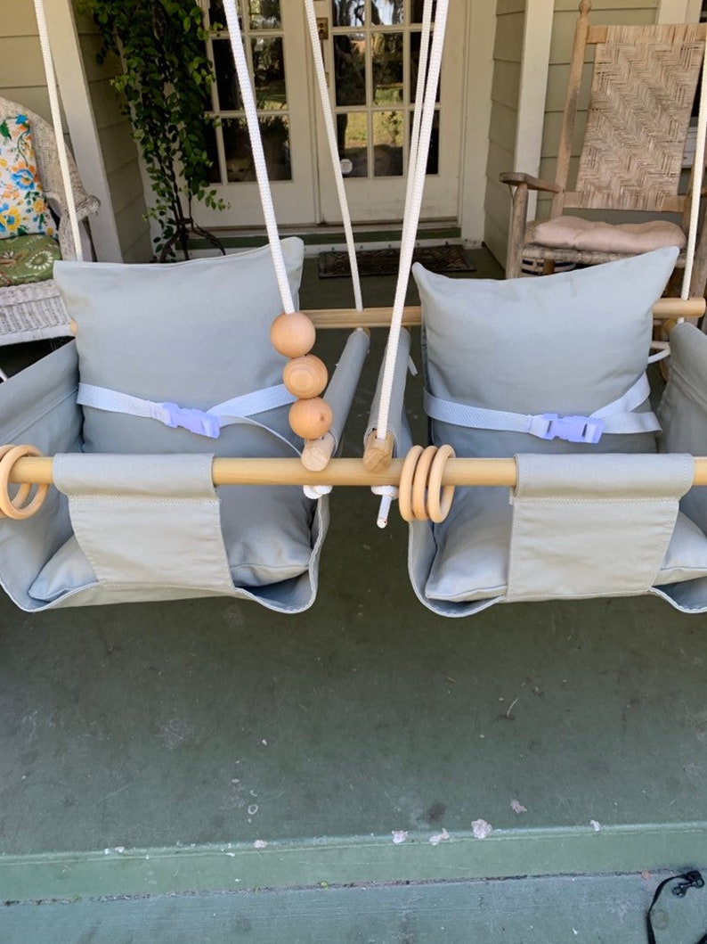 May include: Two gray canvas swing seats with wooden bars and rope hanging straps. Each seat has a gray cushion and a purple buckle strap. There are wooden beads on the bar.