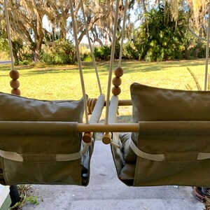 May include: A wooden swing set with two beige fabric seats and wooden bead accents. The swing set is hanging from a porch with a green lawn and trees in the background.