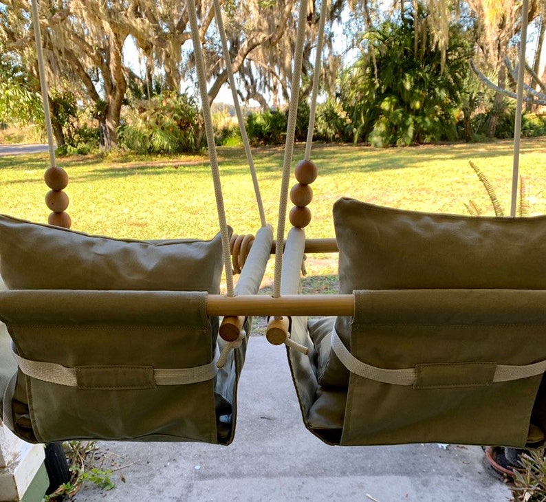 May include: A wooden swing set with two beige fabric seats and wooden bead accents. The swing set is hanging from a porch with a green lawn and trees in the background.