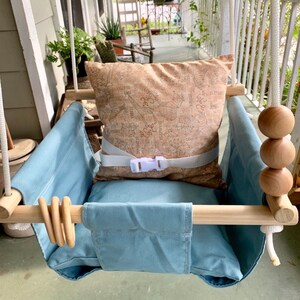 May include: A light blue baby swing with a wooden frame and a brown patterned pillow. The swing has a white rope and wooden beads.