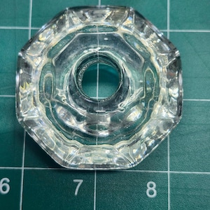 May include: Clear, octagonal glass knob with a central hole. The knob has a faceted design around the edges, reflecting light. The knob is likely for a cabinet or drawer, and is shown against a green surface.
