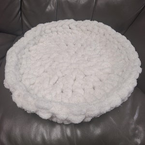 May include: A round, white, knitted pet bed with a textured surface. The bed is made of a soft, fluffy material and is designed for comfort. The bed is circular in shape and sits on a dark gray surface.