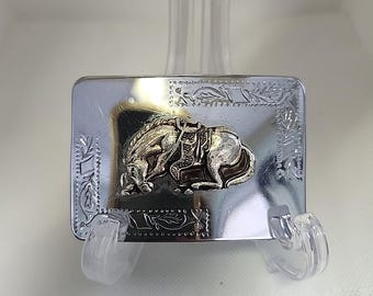 May include: A rectangular silver-colored belt buckle with a detailed relief of a horse. The horse is saddled and appears to be resting. The buckle has an ornate floral border design. The buckle is displayed on a clear stand.