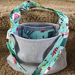 May include: A light blue denim tote bag with a turquoise and white fabric lining. The lining has a pattern of cupcakes, cherries, and polka dots. The bag has a turquoise and white fabric strap with a similar pattern.