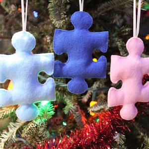 May include: Three felt puzzle piece ornaments hanging on a Christmas tree. The ornaments are blue, light blue, and pink.