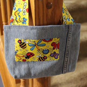 May include: A denim fabric bag featuring a yellow rectangular patch with a colorful insect print including bees, ladybugs, butterflies, and dragonflies. The bag has matching yellow straps with the same insect pattern and the word BUGS printed on them.