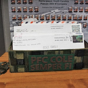 May include: A green and brown camouflage fabric box with a green felt patch that reads "PFC COLE SEMPER FI". The box is filled with white envelopes with handwritten addresses and stamps. The box is sitting on a wooden surface.