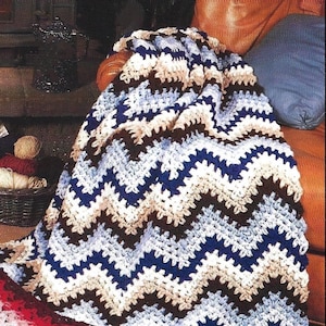 Może przedstawiać: Koc wykonany na szydełku z chevronowym wzorem w odcieniach brązu, niebieskiego i białego. Koc jest zarzucony na krzesło i ma frędzlowany brzeg.