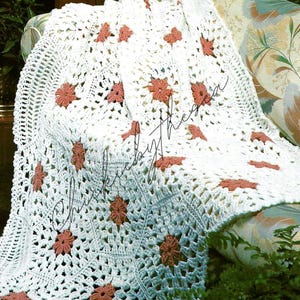 May include: A white crocheted blanket with a floral pattern, draped over a sofa. The blanket features a repeating design of white openwork squares with coral-colored flower motifs. The sofa has floral patterned cushions.