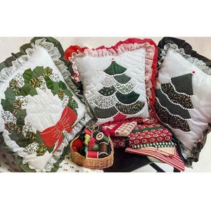 Może przedstawiać: Trzy pikowane poszewki na poduszki z motywami świątecznymi. Jedna przedstawia zielony wieniec z czerwoną kokardką, druga zielone choinki, a trzecia zielone choinki na białym tle. Poduszki są ozdobione białą koronką.