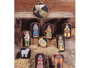May include: A collection of cross-stitch Christmas ornaments depicting the Nativity scene. The set includes figures of Mary, Joseph, the Three Wise Men, an angel, baby Jesus, a sheep, and a camel. Each ornament has a black background with gold trim.