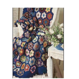 May include: A colorful crocheted throw blanket with a blue background and a floral pattern. The blanket is draped over a white chair and a white table with a vase of white flowers.