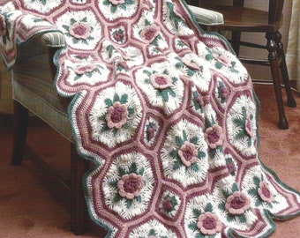 Op de afbeelding: Een gehaakte deken met een bloemenpatroon, gedrapeerd over een stoel. Het patroon van de deken bevat witte, roze en groene elementen, met zeshoekige vormen en roosachtige details. Een decoratief, getextureerd item.