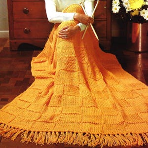 May include: An orange knitted blanket with a fringed edge. The blanket has a textured, square pattern. The person in the image is holding knitting needles and wearing a white turtleneck sweater.