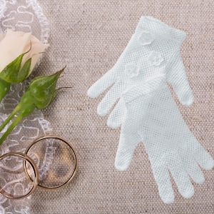 May include: Two gold wedding bands and a white rose with green leaves on a white lace fabric background.  A pair of white lace gloves are on the right side of the image.
