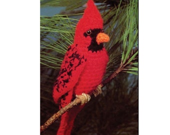 May include: A red cardinal bird with black markings is perched on a branch. The bird is crocheted and has a realistic appearance.