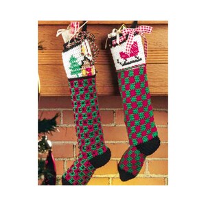 Op de afbeelding: Twee handgemaakte kerstsokken. De sokken hebben een rood en groen geblokt patroon op het lichaam, met een zwarte hiel en teen. De bovenkant van elke sok is wit met een kerstboom en een slee-ontwerp. Elke sok hangt aan een houten schoorsteenmantel.