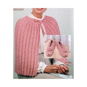 May include: A pink knitted cape with a matching pink ribbon tied around the neck. The cape is shown being worn by a person. The image also shows a pair of pink knitted slippers with a flower detail.