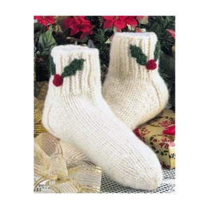 Puede incluir: Un par de calcetines de punto blanco con bordados de acebo verde y bayas rojas. Los calcetines están sobre una superficie con una cinta dorada y una caja de regalo.