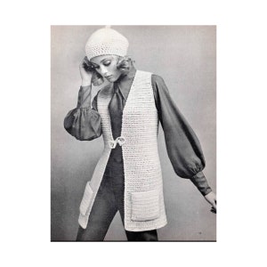 May include: A white crocheted waistcoat with a V-neckline and pockets. The waistcoat has a button fastening and is worn over a black shirt and trousers.