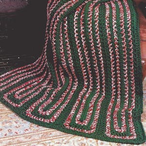 May include: A handmade, green, red, and white crocheted blanket. The blanket features a repeating pattern of vertical green stripes with red and white accents. The blanket is draped over a chair and has a basket of pinecones on top.