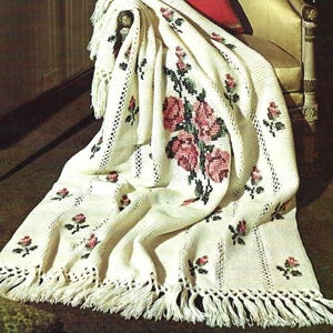 May include: Cream-colored crocheted blanket with a floral rose design in pink and green. The blanket features a fringe along the bottom edge and is draped over a chair. The roses are clustered in the center and scattered across the blanket.
