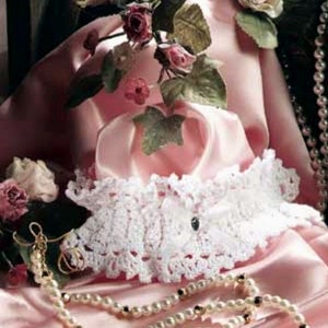 May include: A white crocheted lace garter with a pink satin ribbon and a pearl embellishment. The garter is on a pink satin fabric background with a pearl necklace.
