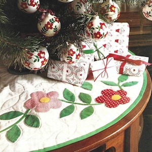 May include: A white quilted table runner with a green border and a floral design. The design features two pink and red flowers with green leaves and stems. The table runner is decorated with a Christmas tree with red and white baubles.