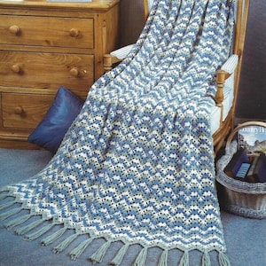 May include: A crocheted throw blanket with a chevron pattern in shades of blue, green, and white. The blanket has a fringe border and is draped over a wooden chair.