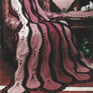 May include: A crocheted throw blanket in shades of pink and black with a lacy pattern. The blanket is draped over a chair and has a scalloped edge.